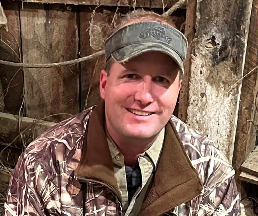 Man in camouflage jacket and cap in rustic outdoor setting.